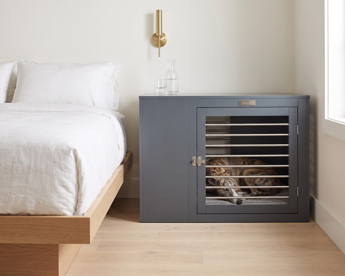 Whippet asleep inside the Moderno Crate in Grey with the doors latched, beside the bed in place of a nightstand